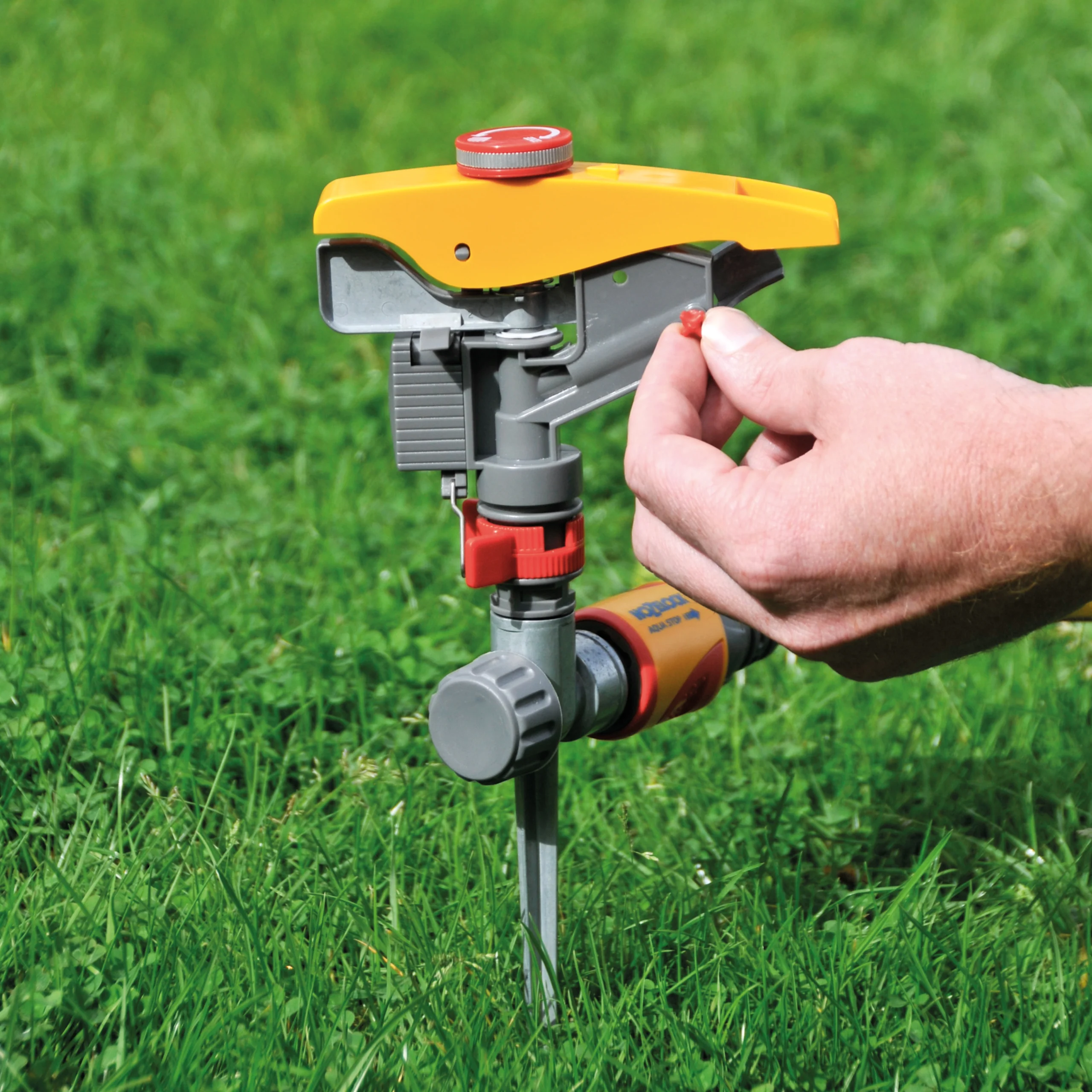 Step 1 for the Hozelock Pulsating Sprinkler controls shown with a hand rotating a red screw with the sprinkler on some grass