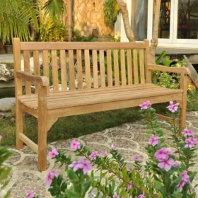 Three-seater teak bench with slatted seat and backrest, placed on a tiled garden path surrounded by flowering plants and greenery, with a house in the background.