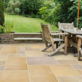 Garden patio with buff sandstone slabs in a mixed-size layout, featuring rustic wooden garden furniture and surrounded by lush greenery and a neatly mowed lawn.