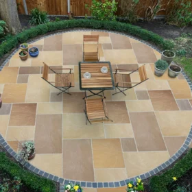 Round garden patio paved with buff sandstone slabs in a mixed-size pattern, bordered with dark cobbles, and furnished with a wooden bistro set surrounded by potted plants and well-kept hedging.