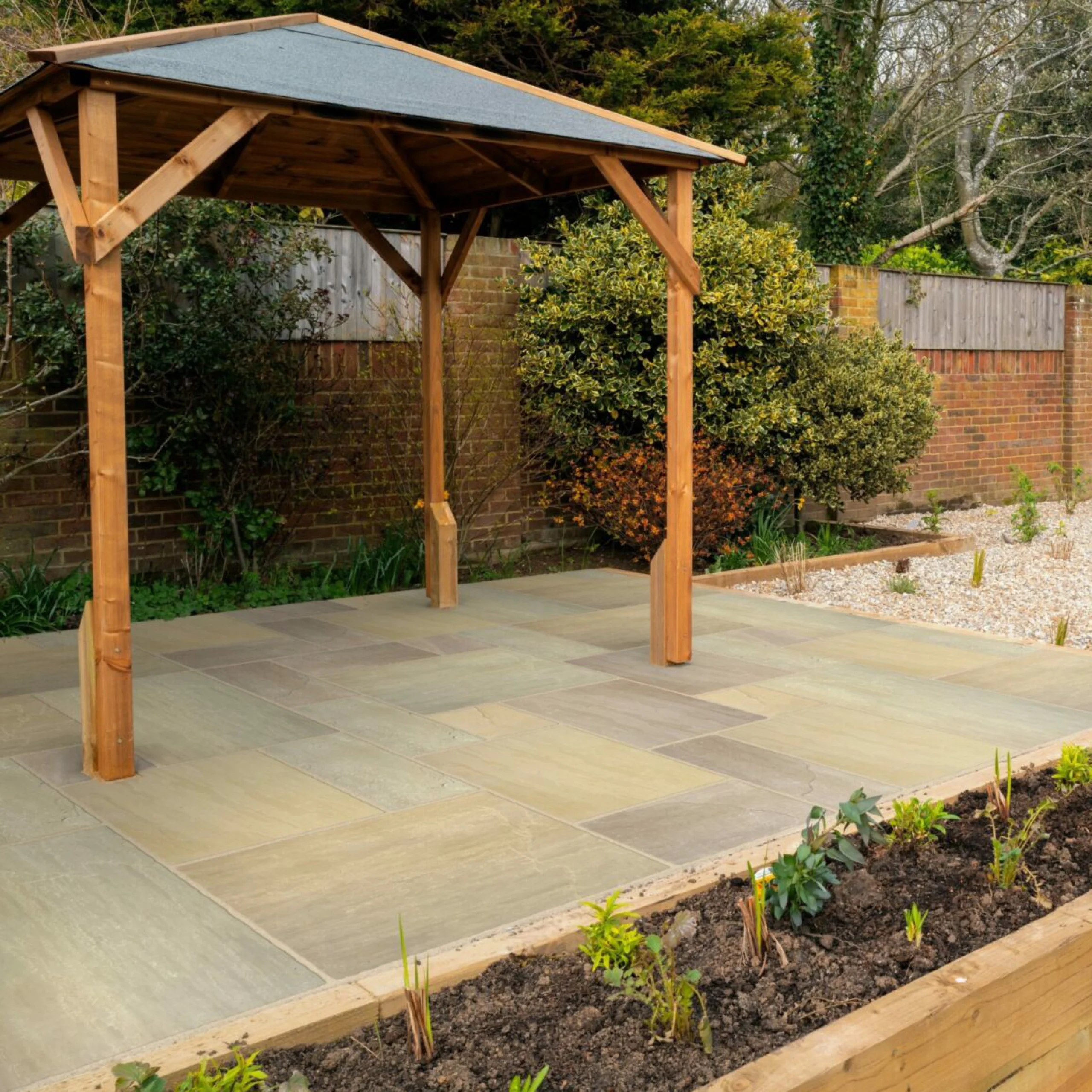 Garden patio area with sandstone Raj Green paving slabs in a mixed-size pattern, featuring a wooden pergola, raised flower beds, gravel sections, and mature border planting against a brick wall backdrop.