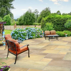 Spacious patio paved with sandstone Raj Green paving slabs in a random pattern, featuring cushioned wrought iron garden furniture, colourful flower pots, and a large border of blooming blue hydrangeas.