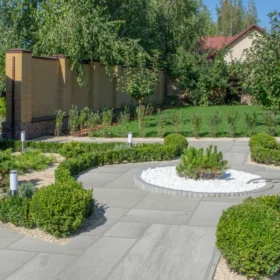 Neatly landscaped garden with Kandla grey sandstone slabs in a random pattern, featuring shaped box hedging, gravel borders, and a circular white gravel bed with a small ornamental conifer.