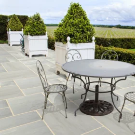 Elegant patio area paved with Kandla grey sandstone slabs in a neat layout, featuring ornate metal garden furniture and white planter boxes with cone-shaped topiary, overlooking a vineyard landscape.