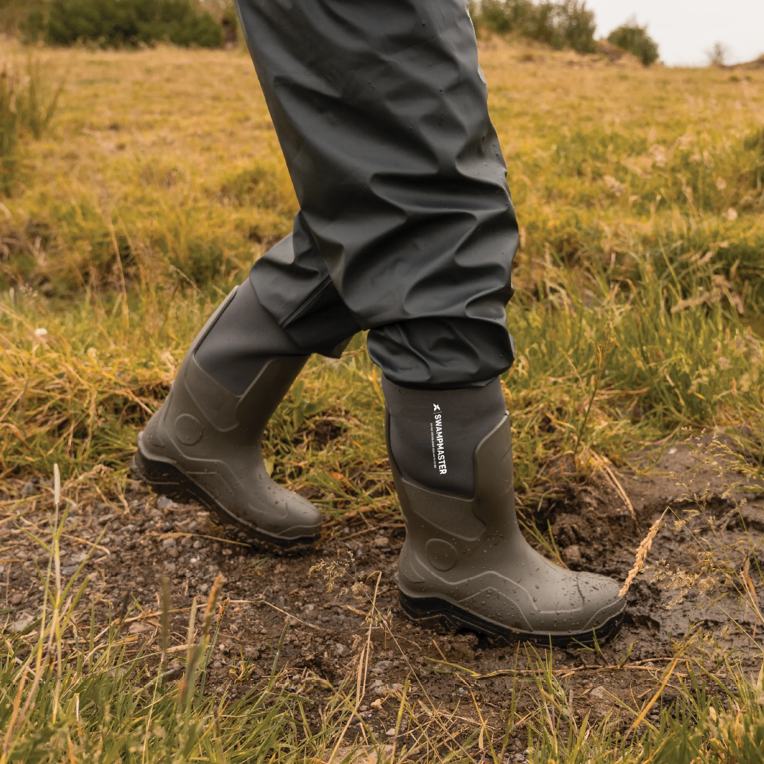A person walking through muddy grass wearing neoprene wellington boots and waterproof trousers, showing the boots in use outdoors.