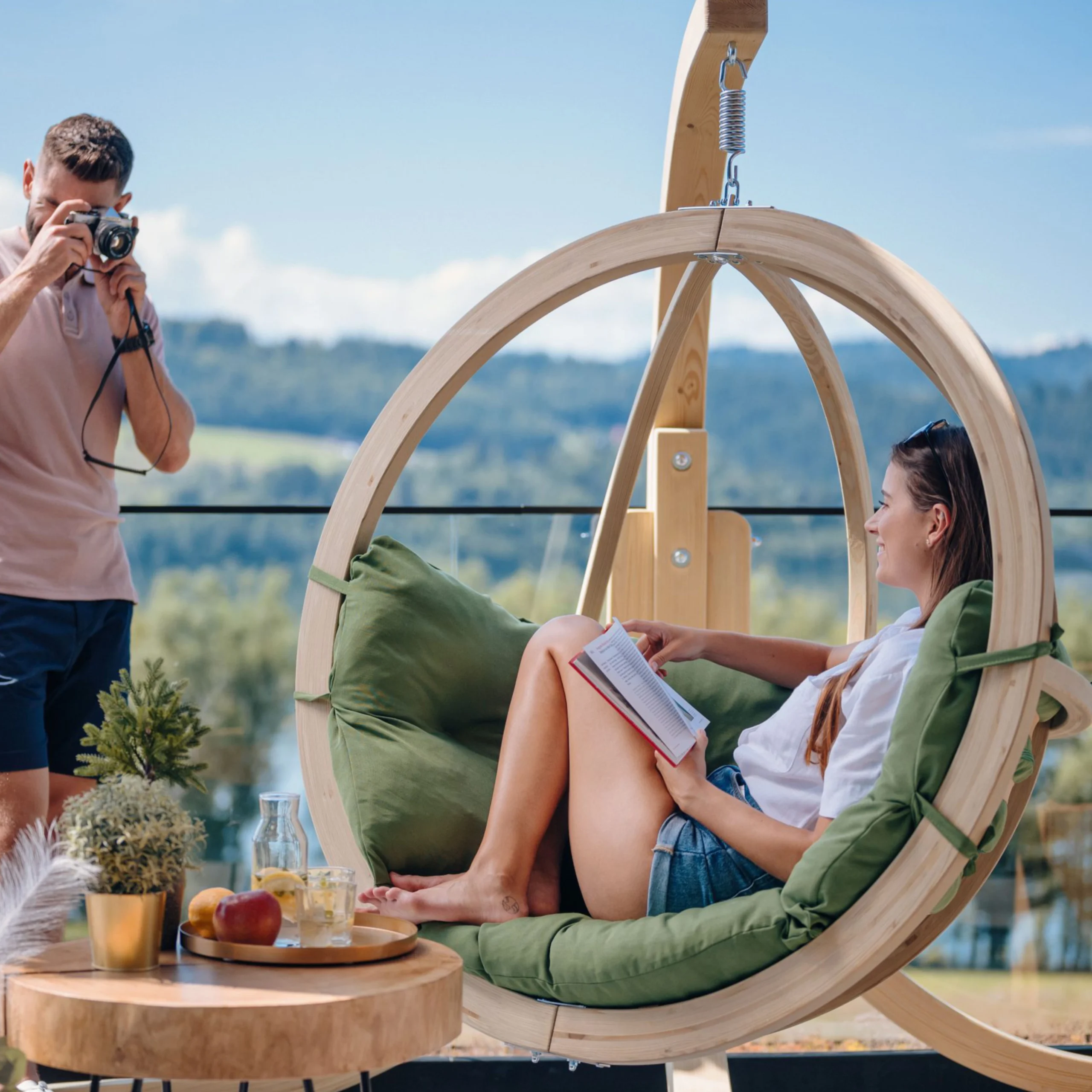 Woman relaxing with a book in a green cushioned egg swing chair on a terrace, showcasing stylish outdoor comfort with a wooden frame design. Garden Swing Seat.
