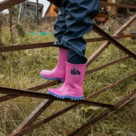 Child wearing Swampmaster pink tractor wellies with lilac soles climbing a farm gate, waterproof PVC kids boots for outdoor adventures.