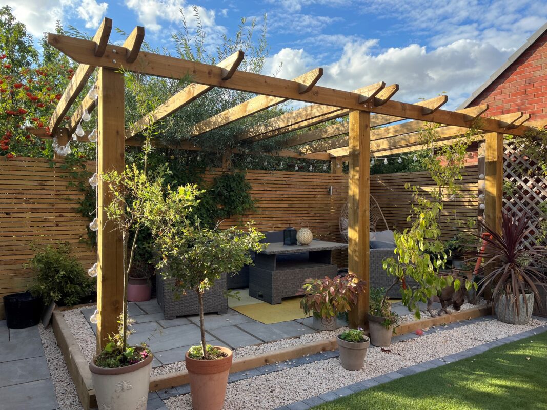 How to build a pergola like this one with six posts and a number of cross beams covering a slabbed garden area complete with table and chairs and decorative potted plants.